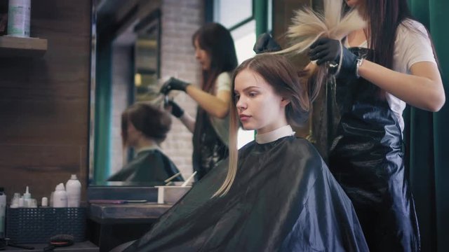 Young blonde woman is sitting in chair in beauty salon while hairdresser is straightening a cape and unties her long hair