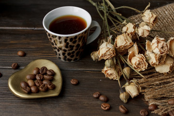 Bronze glass vase with pressed flower on a tablecloth. Golden hearth-shaped saucer with roasted coffee beans. Pure arabica on a wooden table. Lighted candle on the background. Aromatic breakfast