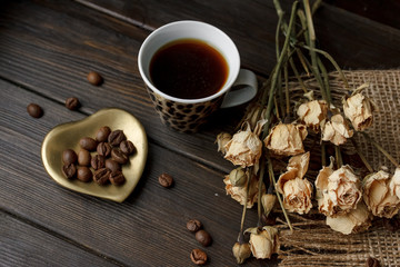 Bronze glass vase with pressed flower on a tablecloth. Golden hearth-shaped saucer with roasted coffee beans. Pure arabica on a wooden table. Lighted candle on the background. Aromatic breakfast