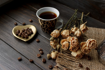 Bronze glass vase with pressed flower on a tablecloth. Golden hearth-shaped saucer with roasted coffee beans. Pure arabica on a wooden table. Lighted candle on the background. Aromatic breakfast