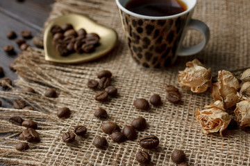 Bronze glass vase with pressed flower on a tablecloth. Golden hearth-shaped saucer with roasted coffee beans. Pure arabica on a wooden table. Lighted candle on the background. Aromatic breakfast