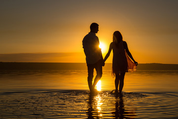 Young couple is walking in the water on summer beach. Sunset over the sea.Two silhouettes against the sun. Just married couple hugging. Romantic love story. Man and woman in holiday honeymoon trip.