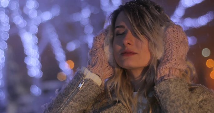 Portrait of a young woman wearing ear muffs with head tilted, trying to warm herself in night winter city. Winter concept. Christmas, winter holidays concept. Snowfall.