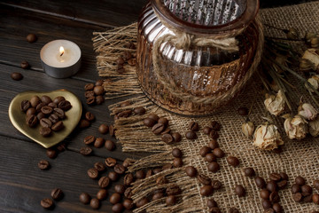 Vase with pressed flower on a tablecloth. Golden hearth-shaped saucer with roasted coffee beans. Pure arabica on a wooden table. Lighted candle on the background. Aromatic breakfast