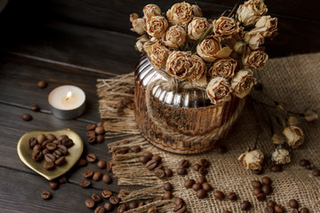 Vase with pressed flower on a tablecloth. Golden hearth-shaped saucer with roasted coffee beans. Pure arabica on a wooden table. Lighted candle on the background. Aromatic breakfast