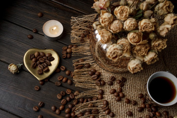 Vase with pressed flower on a tablecloth. Golden hearth-shaped saucer with roasted coffee beans. Pure arabica on a wooden table. Lighted candle on the background. Aromatic breakfast