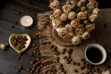 Vase with pressed flower on a tablecloth. Golden hearth-shaped saucer with roasted coffee beans. Pure arabica on a wooden table. Lighted candle on the background. Aromatic breakfast