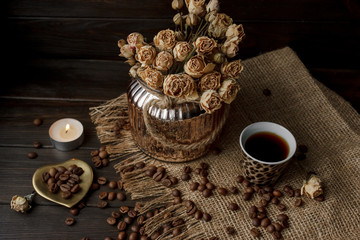Vase with pressed flower on a tablecloth. Golden hearth-shaped saucer with roasted coffee beans. Pure arabica on a wooden table. Lighted candle on the background. Aromatic breakfast