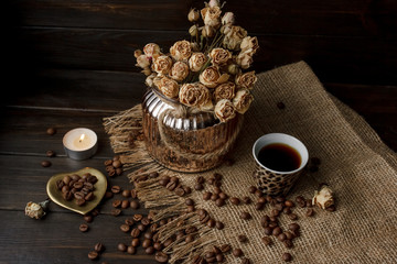 Vase with pressed flower on a tablecloth. Golden hearth-shaped saucer with roasted coffee beans. Pure arabica on a wooden table. Lighted candle on the background. Aromatic breakfast