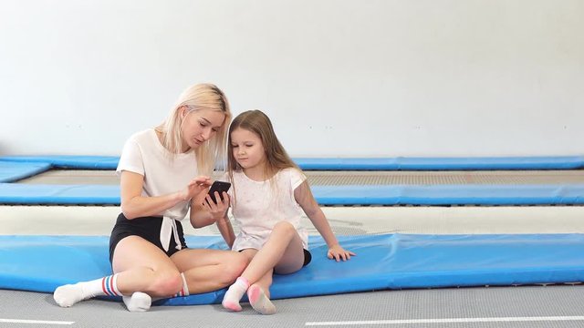 Mother and girl using phone in gym for watching video how do jumping exercise.