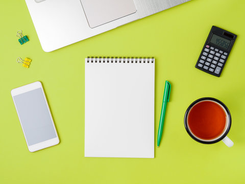 Modern Creative Bright Green Office Desk With Laptop, Smartphone And Other Accessories. Blank Notepad Page For Text In The Middle, Top View.