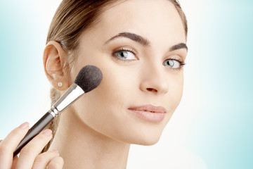 Close-up of a young woman holding brush in her hand and applying makeup. Isolated on light blue background.