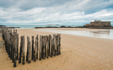 Wooden poles on beach during low tide and Fort National, 17-century fortress on island in background in Saint-Malo, Brittany, France