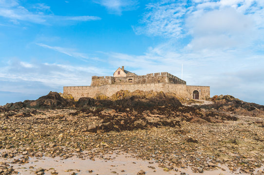 Low angle shot of Fort National, 17-century fortress on tidal island Petit Be surrounded by stones and rocks against bright blue sky in Saint-Malo, Brittany, France