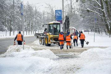 In winter, in snowstorm, workers and a tractor clean the road from snow.