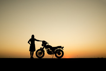Fototapeta premium Silhouette of beautiful women and motorcycles. She standing with her back,beautiful sunset background,Thailand