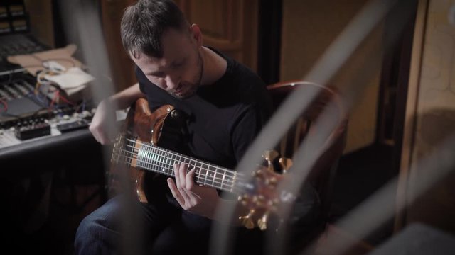 bearded man is plunking electro guitar in dark room of sound recording studio, rehearsing and practicing