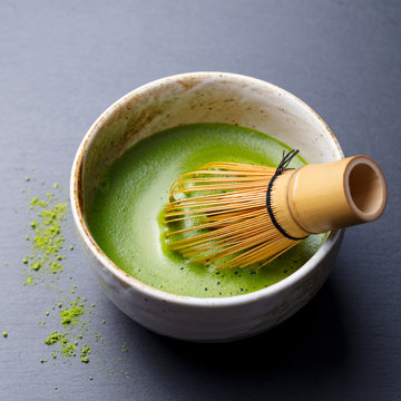 Fototapeta Matcha green tea cooking process in a bowl with bamboo whisk. Black slate background. Close up