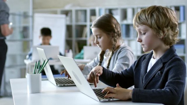 Tracking shot of two primary school children typing something on laptop computer in information technology class, girl trying to copy boy's answers