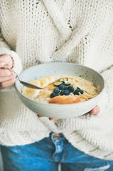 Healthy winter breakfast in bed. Woman in woolen sweater and jeans eating vegan almond milk oatmeal porridge with berries, fruit and almond. Clean eating, vegetarian food concept
