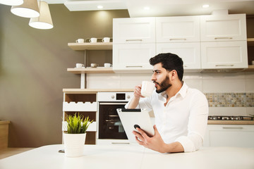 Handsome bearded man with back wavy hair using tablet while having coffee in modern, spacious kitchen, wearing elegant white shirt