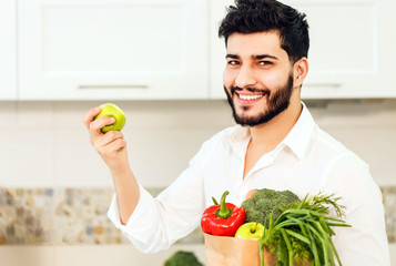 Fototapeta premium Black-haired bearded man, in smart white shirt, holding shopping pack with fresh, organic food and green apple inthe kitchen