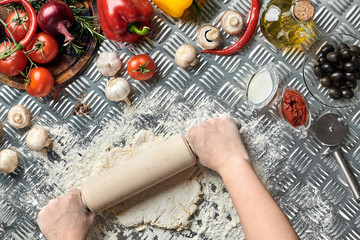 Obraz na płótnie Canvas Female hands roll out dough on metal background, close up. Chef makes dough. Table with vegetables and kitchenware. Italian cuisine concept. Pizza