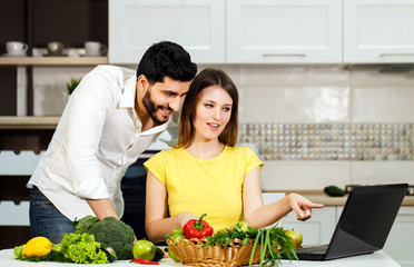 Lovely family couple spending evening together, smiling woman slicing cucumber, happy man standing nearby, using laptop and having pleasant conversation at the table with fresh green vegetables and