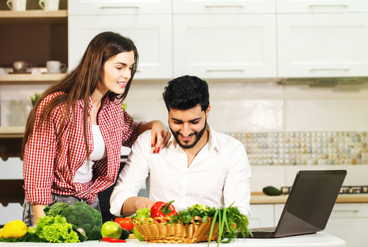 Lovely family couple spending evening together, smiling woman slicing cucumber, happy man standing nearby, using laptop and having pleasant conversation at the table with fresh green vegetables and