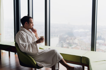 Man in bathrobe drinking coffee at luxury skyscraper hotel near the window with panoramic view. Businessman on a vacation or business trip at the morning