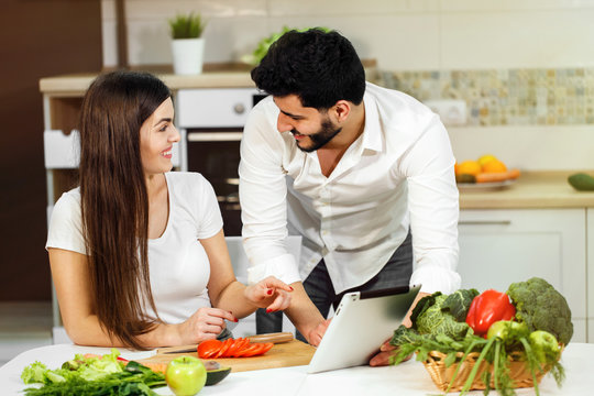 Great family evening, pretty dark-haired woman cutting tomato, handsome tall man showing pictures on tablet, indoor shot in fine white kitchen, concept of healthy eating and beautiful body