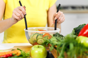 Young woman in yellow t-shirt stirring vegetable salad in a transparent bowl, indoor shot in modern, white kitchen