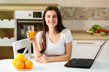 Smiling dark-haired woman having orange juice, positive relaxing atmosphere in kitchen with laptop