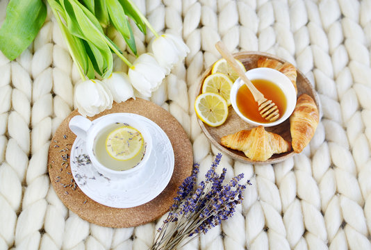 Cup with lavender tea, citrus and honey, croissant, white pastel giant knit blanket, bedroom, flowers tulips, spring, woman day, morning concept