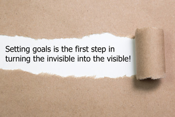 Motivational quote Setting goals is the first step in turning the invisible into the visible , appearing behind torn paper.