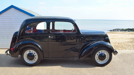 Classic  Black Motor Car parked seafront promenade with sea and beach hut in background.