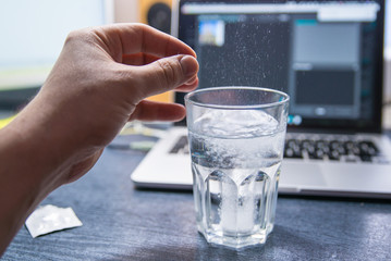 hand through pill of aspirin in glass of water. laptop on background. overloaded with office work