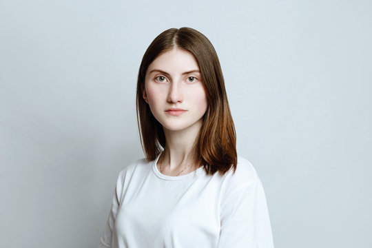 Model portrait without make-up in a white t-shirt on a white background. The girl poses in studio, looking at camera with serious or pensive expression. close up