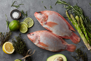 Raw red tilapia fish cooking with herbs, spices, green asparagus, avocado, lemon and lime on gray stone background, top view