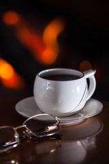 White cup of tea or coffee and glasses near fireplace on wooden table. Winter and Christmas holiday concept