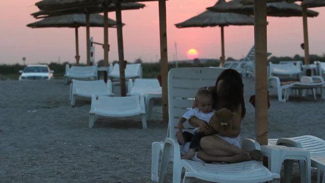 Young mother with baby son sitting on  sunbed play with teddy bear toy, sunset