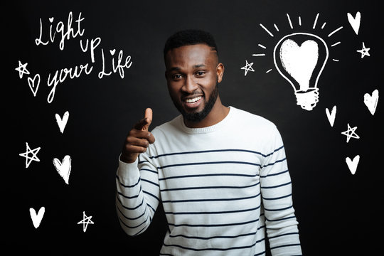 Enjoying life. Cheerful positive young man standing against the gray background and pointing in front of himself while smiling and encouraging other people to enjoy life