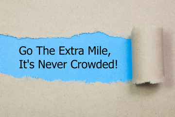 Motivational quote Go The Extra Mile It's Never Crowded appearing behind torn paper.