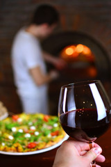 Wineglass with a red wine in a pizzeria restaurant. Chef cooks pizza in a fire stone stove on a background.
