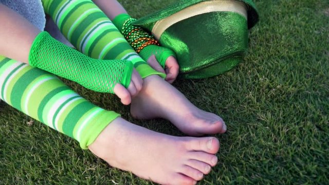 Slow motion. Girl in funny green striped leg warmers fixing it. Saint Patrick's day. Green gloves, hat, bracelets on the wrist. Birthday party. Close up ancle. Vert gants filets. Grass, park. Carnival