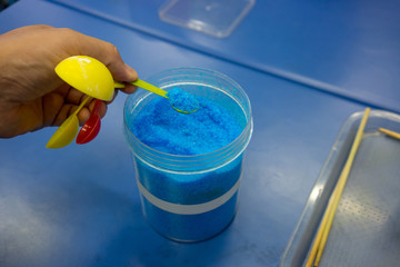 Hands holding spoon of blue chemical, Copper Sulphate Pentahydrate, against blue table