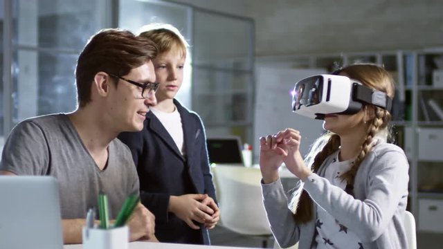 Primary school girl using virtual reality glasses for the first time in information technology class, sitting next to her teacher and classmate