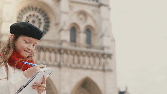 Little stylish girl in beret drawing the picture in notebook near the Notre Dame in Paris, France, doing her hobby.