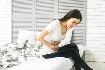 Young woman suffering from abdominal pain while sitting on bed at home. Stomach pain. In stress.
