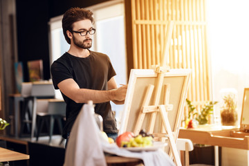 Freelancer man painting with brush standing behind easel.